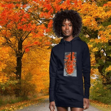 Una mujer con cabello rizado oscuro camina por un parque soleado durante el otoño. Viste una sudadera negra de manga larga que muestra en el centro una reproducción artística de un rostro femenino con trazos abstractos en tonos naranjas. El fondo presenta árboles con hojas doradas y anaranjadas bajo una luz suave.