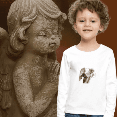 Un niño sonriendo junto a una gran escultura de un ángel, vistiendo una camiseta blanca de manga larga con el diseño de un ángel de la guarda.