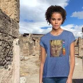Una mujer con cabello rizado posa en las ruinas históricas de Pompeya, Italia, bajo un cielo azul con nubes blancas. Viste una camiseta de color azul jaspeado que presenta en el centro una reproducción artística de un rostro clásico con los ojos cerrados, acompañada de una hilera vertical de círculos de colores que evocan los chakras. A la izquierda se observan antiguos muros de piedra texturizada.
