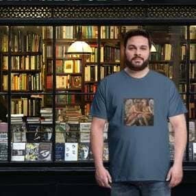 Un hombre frente a una librería antigua iluminada cálidamente. Viste una camiseta de color azul petróleo que muestra en el centro una reproducción artística de dos figuras clásicas o ángeles entrelazados en tonos tierra. A través del cristal se aprecian estanterías repletas de libros y una lámpara de estilo clásico.